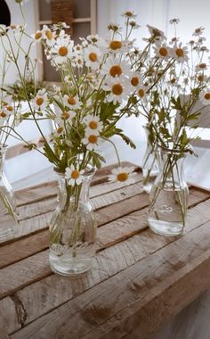 three clear vases filled with white and yellow daisies on a wooden table in front of a window