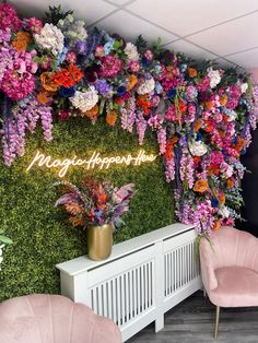 a room with pink chairs and purple flowers on the wall, along with a neon sign that reads magic happens