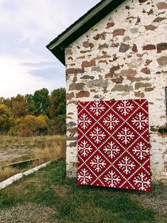 a red and white quilt hanging on the side of a brick building next to a field