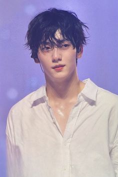 a young man with black hair and white shirt looking off to the side while standing in front of a purple background