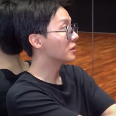 a man with black hair and glasses sitting in front of a mirror on a wooden floor