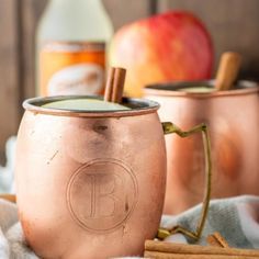 two copper mugs sitting on top of a table next to cinnamon sticks and an apple