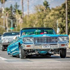 an old blue car driving down the road with other cars behind it and palm trees in the background