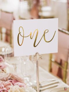 a close up of a sign on a table with flowers in the foreground and an empty wine glass behind it