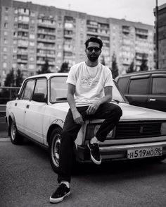 a man sitting on the hood of a car in front of some buildings and parked cars