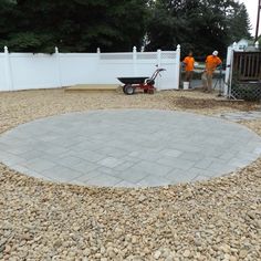 two men in orange shirts are standing around a circular stone area with a wheelbarrow