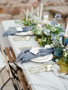 the table is set with white plates and silverware, blue napkins and flowers