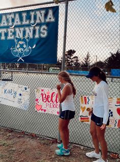 two girls standing next to each other in front of a fence with signs on it