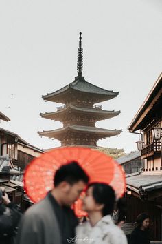 two people standing in front of a tall building with an orange umbrella over their heads