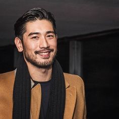 a man with a beard wearing a brown jacket and black scarf smiles at the camera