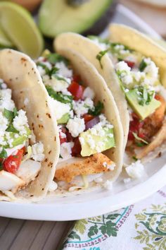 three fish tacos on a plate with avocado and feta in the background