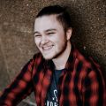 a man smiling while leaning against a wall with his hands on his hips and wearing a black and red flannel shirt