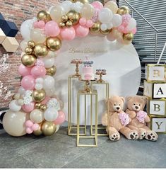 a teddy bear sitting in front of a balloon arch with pink, white and gold balloons