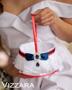 a woman in white dress holding a red and blue wedding garter with diamond brooch on it