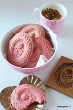 a bowl filled with pink cookies next to a box of chocolates and a cup of coffee