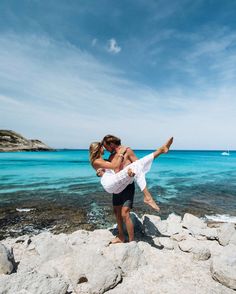 a man and woman are standing on rocks by the ocean with their arms around each other