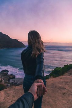 two people holding hands while standing on top of a hill near the ocean at sunset