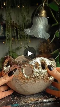 two hands holding up a gold and silver object in front of a planter filled with potted plants