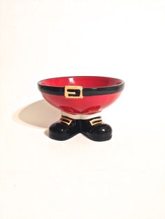 a red bowl with black feet and a gold buckle on the top is sitting in front of a white wall