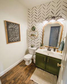 a bathroom with wood flooring and white walls has a green cabinet, mirror, toilet and chalkboard on the wall