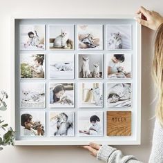 a woman holding up a white framed photo with many pictures on the wall behind her