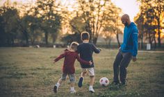 three young boys playing with soccer balls in a park at sunset or sunrise, while an adult looks on