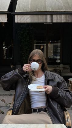 a woman sitting in a chair drinking from a white coffee cup while holding a plate with food on it