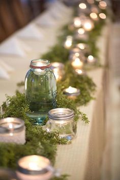 candles are lined up on a table with moss and glass jars in front of them