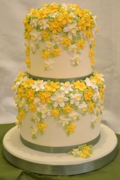 a three tiered cake with yellow and white flowers on the top, sitting on a green tablecloth