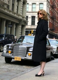 a woman is standing on the sidewalk in front of a black car and some buildings