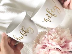 two white cups with gold lettering are next to a pink flower on a white blanket