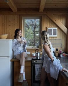 two women sitting on the counter in a cabin kitchen, one drinking from a mug