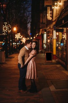 a man and woman standing next to each other on a sidewalk at night with lights in the background