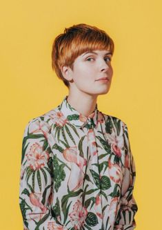 a woman with short red hair wearing a floral shirt and jeans, standing in front of a yellow background