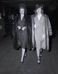 two women dressed in coats and hats walking down a street together, one is wearing a fur coat