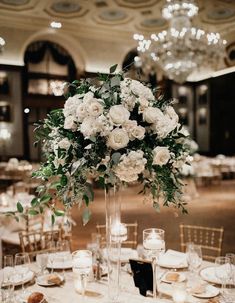 a tall vase filled with white flowers and greenery on top of a round table