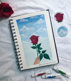 an open notebook with some markers and pens on it next to a drawing of a rose