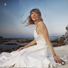 a woman in a white dress is sitting on the beach with birds flying around her