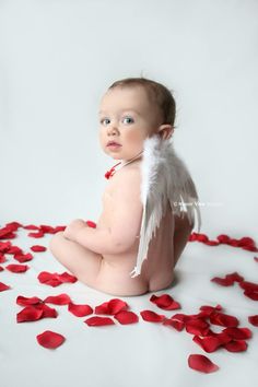 a baby sitting on the ground surrounded by rose petals with an angel's wing