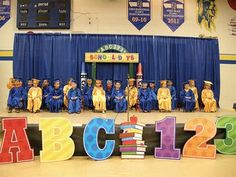 a group of children in graduation gowns sit on the stage with letters spelling out abc and 123