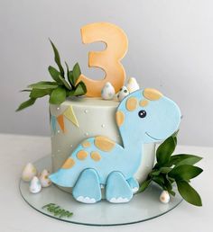 a blue dinosaur cake with the number three on it's top and green leaves