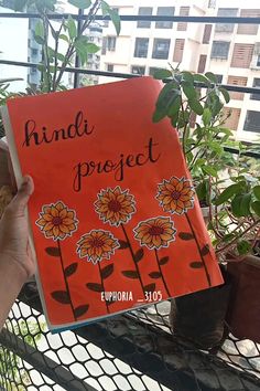 a person holding up a book in front of a fence with plants and buildings behind it