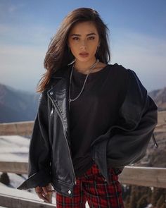 a woman in black jacket and plaid pants standing on a balcony with mountains behind her