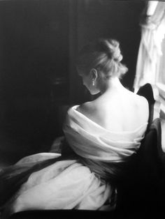 a black and white photo of a woman in a dress sitting on a chair looking out the window