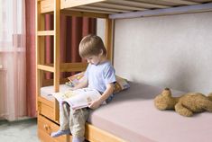 a young boy sitting on top of a bunk bed reading a book next to a teddy bear