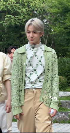 a young man with blonde hair wearing a green jacket and tan pants standing in front of trees