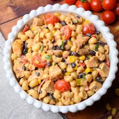 a white bowl filled with pasta salad next to tomatoes and corn on a wooden table