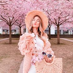 a woman wearing a pink hat and holding a straw bag in front of cherry blossom trees