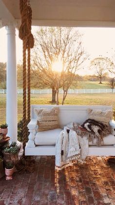 a white porch swing with pillows and blankets on it, in front of a tree