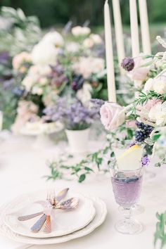 the table is set with white and purple flowers in vases, candles, and plates